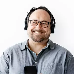  Imagem de um homem branco, sorrindo, com cabelo curto usando óculos e fone de ouvido, sobre um fundo branco